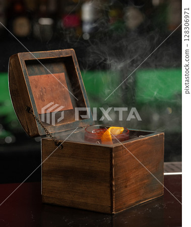 A smoky, dark red alcoholic cocktail with ice and an orange peel garnish is presented in a rocks glass nestled inside an open wooden box. 128306971