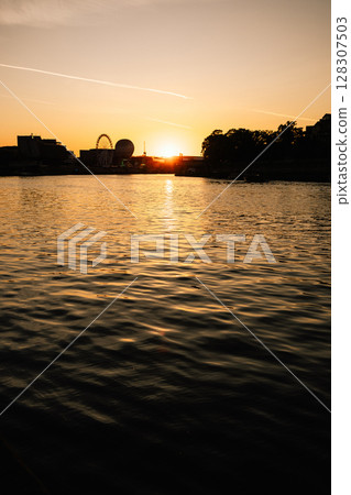 Vertical screen: shadowed riverside view with distant silhouette of Ferris wheel and urban buildings reflecting on water surface, capturing moody atmosphere and gentle evening light. Summer City Mood 128307503