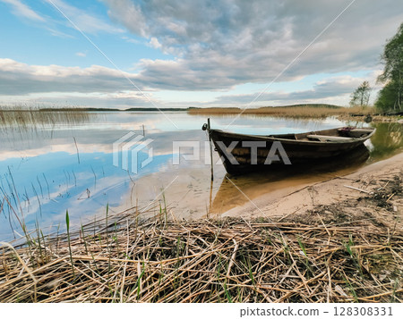 Old wooden fishing boat is moored on shore of calm lake, reflecting blue and cloudy sky, surrounded by reeds and dry grass, creating peaceful and evocative scene. Wooden fishing boat rests on lakeside Old wooden fishing boat is moored on shore of calm lake, reflecting blue and cloudy sky, surrounded by reeds and dry grass, creating peaceful and evocative scene. Wooden fishing boat rests on lakeside 128308331