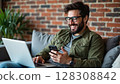 A smiling man wearing glasses relaxes on a sofa, simultaneously using a laptop and a smartphone with a brick wall background. - Generic AI Image 128308842