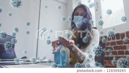 Image of covid 19 cells floating over biracial woman wearing face mask disinfecting her hands in off 128309265