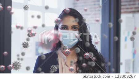 Image of covid 19 cells floating over portrait of biracial woman wearing face mask in office 128309267