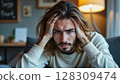 Portrait of a frustrated young man holding his head with both hands, sitting indoors on a couch  Generic AI Image 128309474