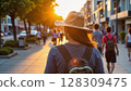 Back view of a woman wearing a hat and backpack walking on a sunny street with people in the background - Generic AI Image 128309475
