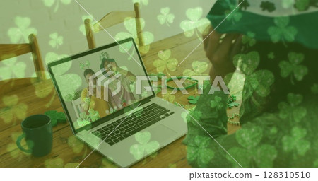 Clover leaves falling against woman wearing shamrock glasses while having a image call at home 128310510