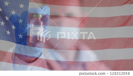 Image of american flag waving over male doctor in face mask holding covid 19 vaccine 128312058