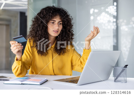 Woman sitting at desk holding credit card while looking at laptop with frustrated expression. Indicates online payment issue or possible fraud, confusion over transaction or account error. Woman sitting at desk holding credit card while looking at laptop with frustrated expression. Indicates online payment issue or possible fraud, confusion over transaction or account error. 128314616