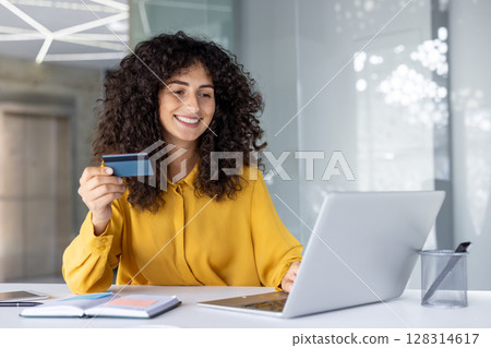 Smiling woman holding credit card using laptop for online shopping or financial transactions. Professional atmosphere in modern office setting, conveying ease and convenience of digital banking Smiling woman holding credit card using laptop for online shopping or financial transactions. Professional atmosphere in modern office setting, conveying ease and convenience of digital banking 128314617
