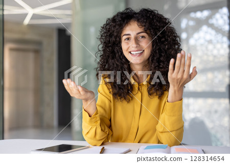 A cheerful woman with curly hair, gestures expressively while smiling, possibly presenting or explaining something in an office setting. 128314654