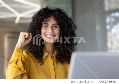A cheerful woman with curly hair smiles with a fist raised, exuding success and confidence. She wears a yellow shirt. 128314659