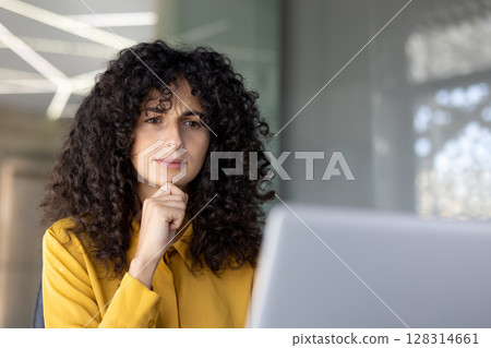 A woman with curly hair looks thoughtfully at a laptop, resting her chin on her hand. 128314661