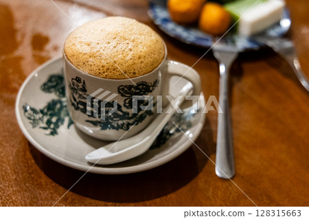 Traditional aromatic frothy coffee with milk served in retro cup and saucer paied with sweet kuih muih at kopitiam in Malaysia Traditional aromatic frothy coffee with milk served in retro cup and saucer paied with sweet kuih muih at kopitiam in Malaysia 128315663