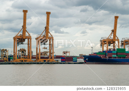 Shipping cranes work in a busy port, lifting cargo containers against a cloudy sky. A vital hub of trade and transport. 128315935