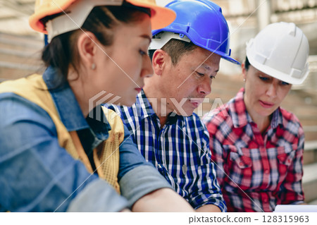 Three construction workers collaborating on project plans at a job site with hard hats and focused expressions. 128315963