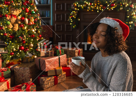 Merry Christmas. African American woman in Santa hat with cup of hot drink coffee tea near Christmas tree in dark interior. Girl in living room with Christmas tree and fireplace. Christmas eve at home Merry Christmas. African American woman in Santa hat with cup of hot drink coffee tea near Christmas tree in dark interior. Girl in living room with Christmas tree and fireplace. Christmas eve at home 128316685