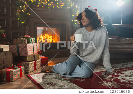 Merry Christmas. African American woman in Santa hat with cup of hot drink coffee tea near Christmas tree in dark interior. Girl in living room with Christmas tree and fireplace. Christmas eve at home Merry Christmas. African American woman in Santa hat with cup of hot drink coffee tea near Christmas tree in dark interior. Girl in living room with Christmas tree and fireplace. Christmas eve at home 128316686
