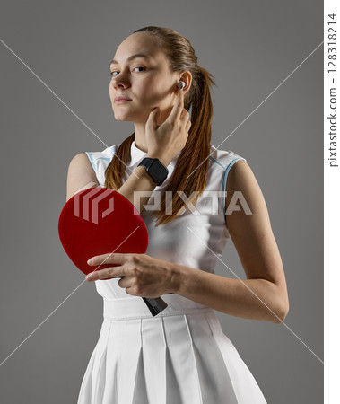 Focused female table tennis player adjusting earbud while holding paddle on gray background 128318214