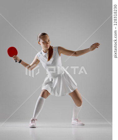 Focused female table tennis player in white outfit preparing to strike on gray background Focused female table tennis player in white outfit preparing to strike on gray background 128318230