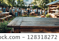 Rustic wooden table in vibrant outdoor setting, surrounded by lush greenery, blurred crowd enjoying a sunny day, creating a warm 128318272