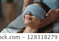 Woman receiving a relaxing facial massage with a soothing eye mask, enjoying a tranquil spa environment, surrounded by soft textures and calming colors, promoting wellness and self-care 128318279