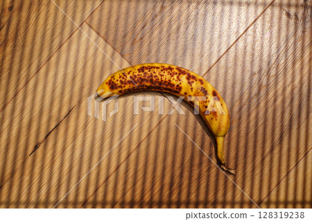 Brown-spotted banana resting on wooden surface with striped shadows during afternoon light 128319238
