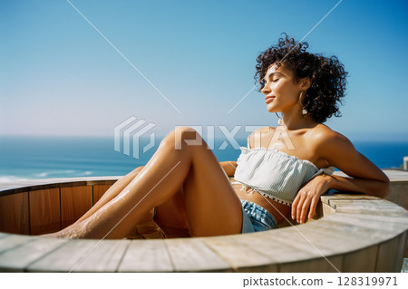 A woman is sitting in a small wooden tub by the ocean 128319971