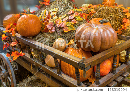 An autumn arrangement featuring various types of pumpkins, straw, and leaves on rustic wooden cart 128319994