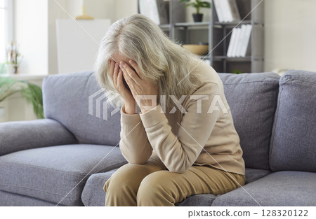Old senior woman sitting indoors on sofa in distress at home, covering face with her hands, weeping  128320122