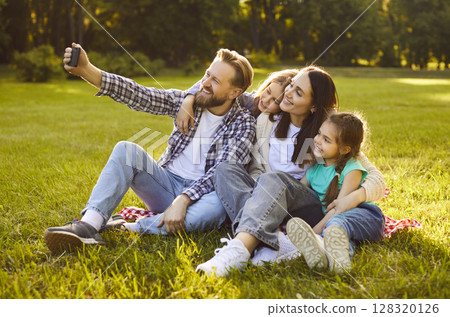 Happy family sitting together on green summer grass taking selfie using mobile phone hugging sharing 128320126