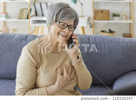 Senior smiling woman pensioner sitting on sofa in living room at home and talking by smartphone. 128320147