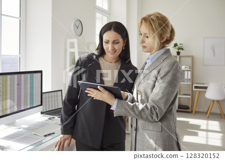 Two businesswomen standing in office with tablet, discussing financial report and analyzing data. 128320152