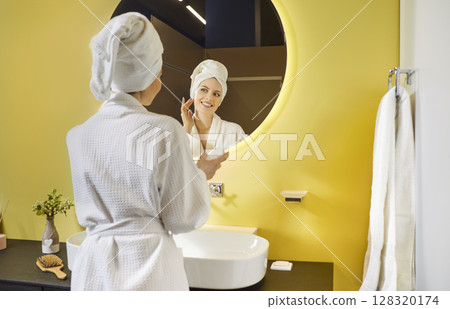 Young woman in bathrobe applying moisturizer while looking in mirror during morning beauty routine. Young woman in bathrobe applying moisturizer while looking in mirror during morning beauty routine. 128320174