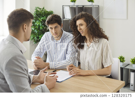 Young family couple sitting at desk, listening to real estate agent during consultation in office. Young family couple sitting at desk, listening to real estate agent during consultation in office. 128320175