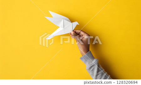 Hand holds paper dove against bright yellow background, representing creativity, peace, childhood and origami art. Hand holds paper dove against bright yellow background, representing creativity, peace, childhood and origami art. 128320694