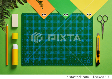 Top view of art workplace in green and yellow colors. Craft knife, pencil, eraser, glue, cutting mat, colored paper. Space for your text. 128320994
