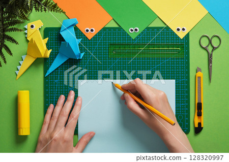 Making an origami dinosaur. The hand draws a pattern for cutting out on paper. Around are items for paper crafts. 128320997