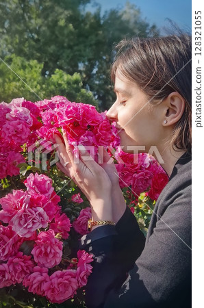 Lovely young woman enjoys the presence in the rose garden as enjoys smelling a large cluster of vibrant pink rosy, savouring the fragrance with closed eyes, gently touching the blooms 128321055