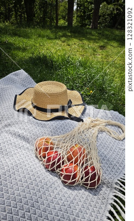 Straw hat and eco mesh bag with peaches on picnic blanket in sunny green meadow 128321092