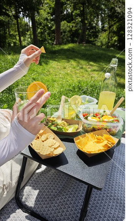 Hand holding glass with orange slice and snacks on picnic table with salad and lemonade in green park 128321094