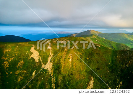 Breathtaking aerial view of green, rugged mountain peaks under dramatic sky. Scene captures contrast between vibrant landscape and moody clouds, with sunlight breaking through. Carpathians, Chornogora 128324362