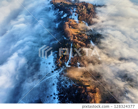 Aerial view of mist-covered landscape where sunlight penetrates through clouds, revealing mix of land and dense forest. Shadows of trees and mist create dramatic interplay of light and dark. 128324377