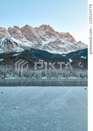 Picturesque Alpine Ski Resort Nestled between Frozen Lake and Snow Covered Mountain Peaks with Traditional Wooden Chalets Reflecting in Crystal Clear Water Surface during Winter Season in European 128324779