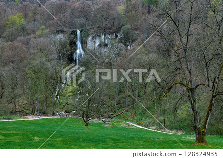 Lush Green Meadow With Cascading Waterfall And Winding Dirt Path Amidst Early Spring Forest Tranquility. Lush Green Meadow With Cascading Waterfall And Winding Dirt Path Amidst Early Spring Forest Tranquility. 128324935