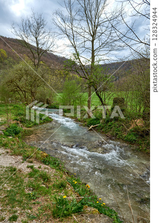Tranquil Spring River Landscape With Lush Green Canopy And Gravel Path Along Serene Flowing Water Amidst Rolling Hills. 128324944