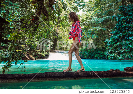 Attractive woman in bikini with a plaid shirt on a log over a turquoise river in the jungle with a waterfall. Tropical holiday adventure. 128325845
