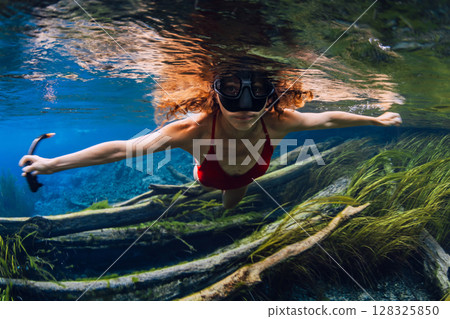 Woman in mask swims underwater over sunken logs with algae in crystal-clear fresh water lake 128325850