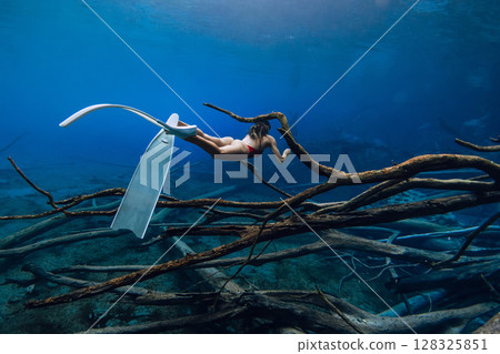 Freediver woman glides underwater with diving fins over sunken logs in the lake. Free diving in blue crystal lake 128325851