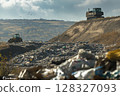 A massive pile of trash with plastic bags and waste looms at the landfill, while bulldozers operate in the background against a cloudy sky. 128327093