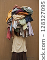 Individual stands overwhelmed by a towering pile of laundry, humorously hiding their face, set against a simple brown backdrop. 128327095