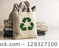 Cotton tote bag featuring a green recycling symbol is filled and placed beside neatly stacked sweaters in a tidy office environment. 128327100